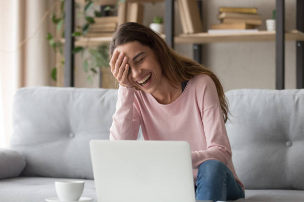 donna ridendo seduta sul divano a guardare film comici su laptop - ridere foto e immagini stock