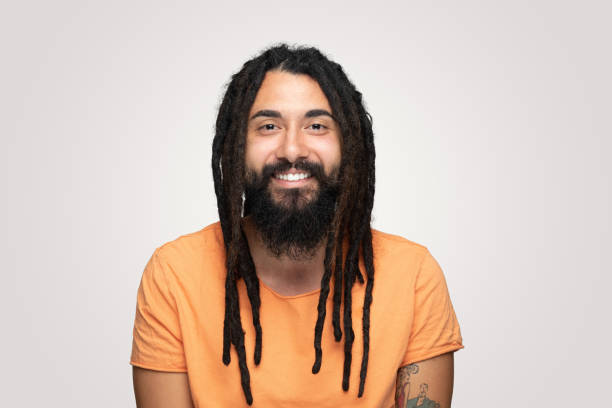 Laughing young handsome bearded man hippie with dreadlocks over isolated  gray background Laughing young handsome bearded man hippie with dreadlocks over isolated  gray background.He standing on isolated background and smile to the camera.Studio shot, isolated background,horizontal composition.Young man wearing orange tshirt and he has long and black hair. big smile emoji stock pictures, royalty-free photos & images