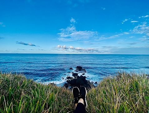 Horizontal vanishing point high up seascape view of ocean with cliff rocks from laying on grass looking out to open horizon over water with crossed feet at Ballina headland NSW Australia