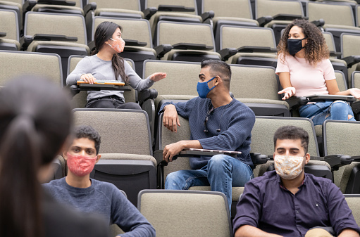 College/university students are seated one seat apart from each other to maintain social distancing. They are all wearing their face mask to protect themselves and others from germs. Some students appear to be chatting with one another while others are looking at their professor.