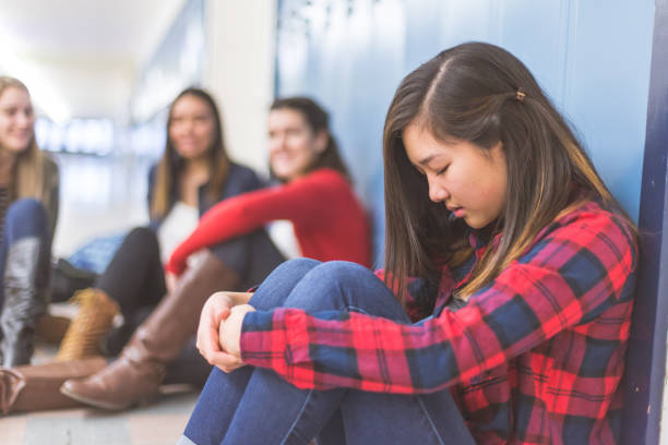 Left Out A sad and left-out ethnic high school student leans against a panel of lockers with her eyes closed, head down, and hands clasped to her knees. Three girls are in the background talking amongst themselves and laughing at her. bullying stock pictures, royalty-free photos & images