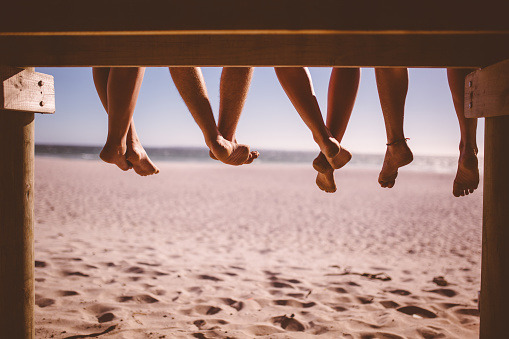 Rearview cropped shot of a row of legs of friends sitting who are sitting in a row on a beach boardwalk with sand and sea in front of them