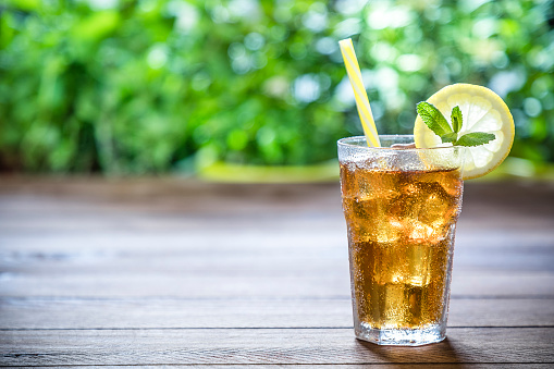 Front view of a drinking glass full of ice cubes and lemon ice tea. The dinking glass is wet with condensed water and has a yellow dinking straw, a lemon slice and a mint leaf on top and is on a rustic wooden table. The main focus is on the drinking glass and the green background is defocused giving a feeling of freshness. The Objects are at the right side of the image leaving a useful copy space at the left side of the glass. Low key DSLR photo taken with Canon EOS 6D Mark II and Canon EF 24-105 mm f/4L