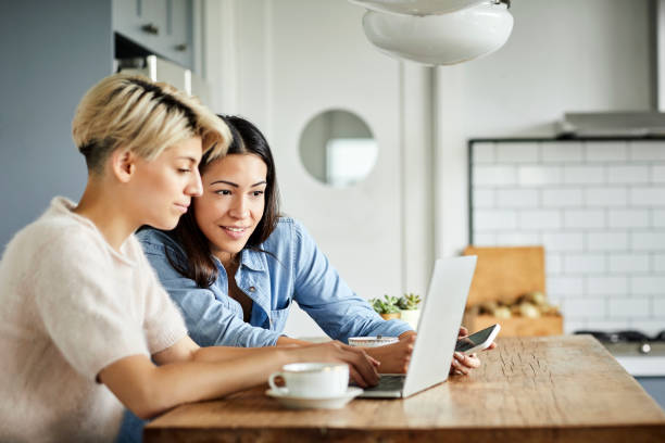 Lesbian couple with laptop on table at home Smiling woman looking at girlfriend using laptop. Lesbian couple is sitting at table in kitchen. They are at home. laptop couple stock pictures, royalty-free photos & images
