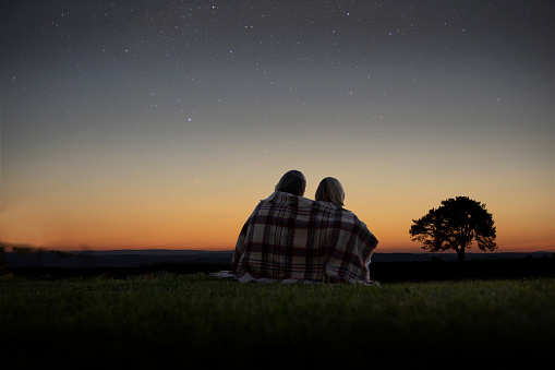 A mature couple sitting wrapped in a blanket looking at the night sky full of stars.