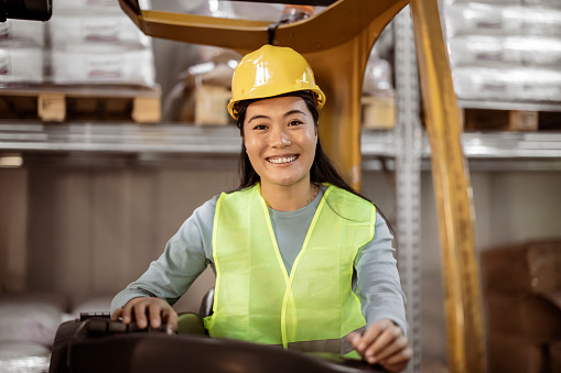 Asian Female forklift driver working in a warehouse. Portrait of Mongolian female forklift operator in safety vest and hardhat looking at camera and smiling