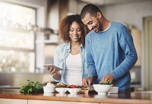 Let's give this recipe a try Shot of a happy young couple using a digital tablet while preparing a healthy meal together at home healthy dinner stock pictures, royalty-free photos & images
