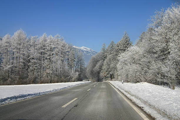 A road through a wooded area in the winter. Mountains visible at the...