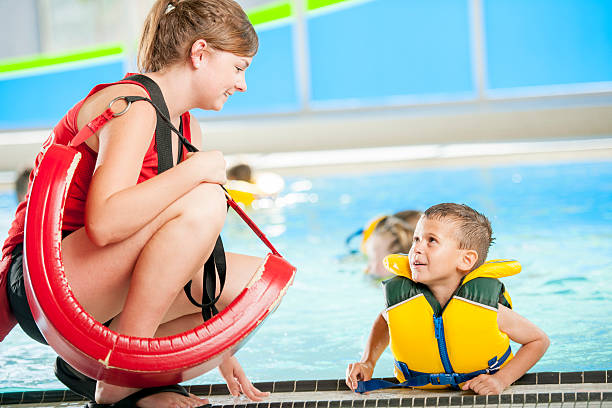 Lifeguard Talking to Child A lifeguard and swim instructor talking to a young boy who is wearing a lifejacket in the pool with other children during a swim class. lifeguard stock pictures, royalty-free photos & images