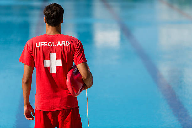 Lifeguard watching swimming pool Rear view of male lifeguard with emergency equipment in red uniform watching swimming pool. lifeguard stock pictures, royalty-free photos & images