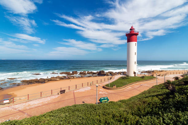 Lighthouse Against Blue Cloudy Coastal Seascape in South Africa Paved Green vegetation and patterned walkway with metal barrier against red and white lighthouse against blue cloudy coastal seascape at Umhlanga, Durban, South Africa durban stock pictures, royalty-free photos & images