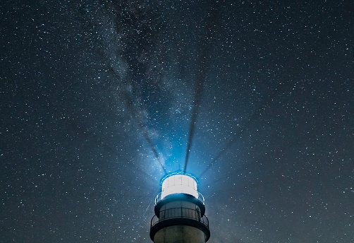 Top part of New England lighthouse isolated against dark, starry sky, with light beams radiating upward