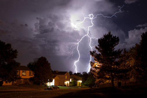 A massive thunderhead pours rain and lightning over suburban Denver homes, Colorado. Trees are silhouetted and homes are lit by incandescent light. The thunderhead is lit by huge lightning bolts reaching from the top of the clouds to the ground.