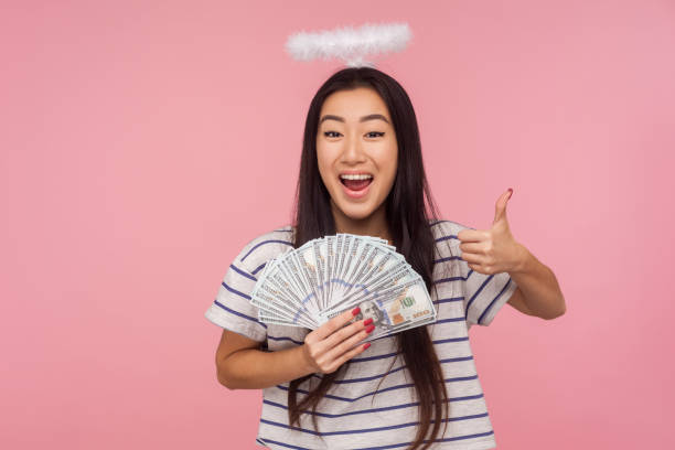 I like my rich life! Portrait of joyful angelic girl with nimbus over head showing thumbs up and holding money I like my rich life! Portrait of joyful angelic girl with nimbus over head showing thumbs up and holding money, satisfied with big income, lottery winning. studio shot isolated on pink background bills saints stock pictures, royalty-free photos & images