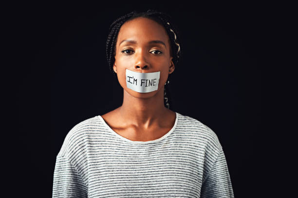 Listen to what's not being said Studio shot of a young woman with a label saying “I’m fine” covering her mouth against a black background prejudice stock pictures, royalty-free photos & images