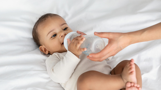 Feeding And Childcare Concept. Closeup of adorable little African American infant wants to drink milk or water, woman holding bottle in hands, feeding her baby who lying on the back on white bedsheets