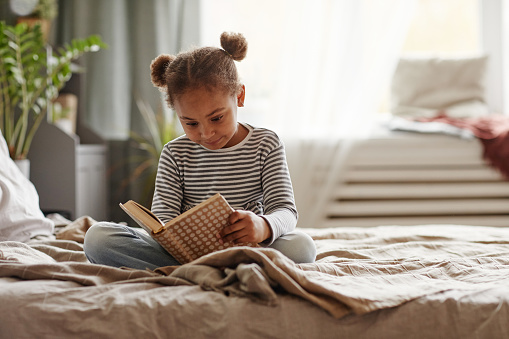 Full length portrait of cute African-American girl reading book while sitting on bed in cozy interior, copy space