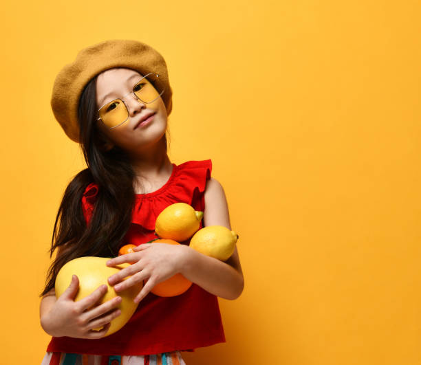 Little asian girl in sunglasses, brown beret, red blouse. Excited, holding pomelo, oranges and lemons. Studio shot
