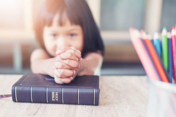a menina asiática pequena pray com a bíblia na sala de aula na escola, conceito do estudo da bíblia - catolicismo - fotografias e filmes do acervo