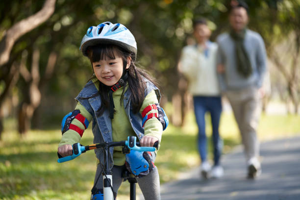 little asian girl riding bike outdoors in park parents watching from behind stock photo