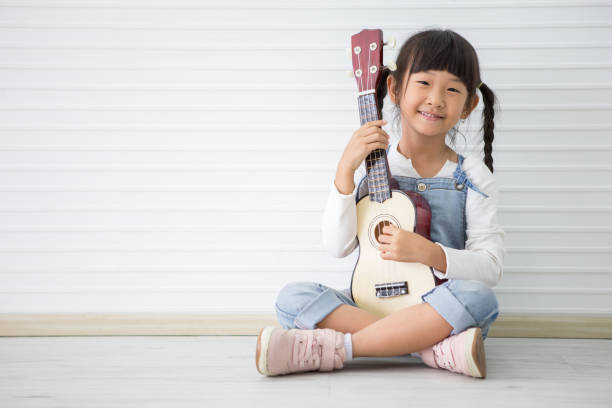 little asian girl sitting playing ukulele on white background with copy space stock photo