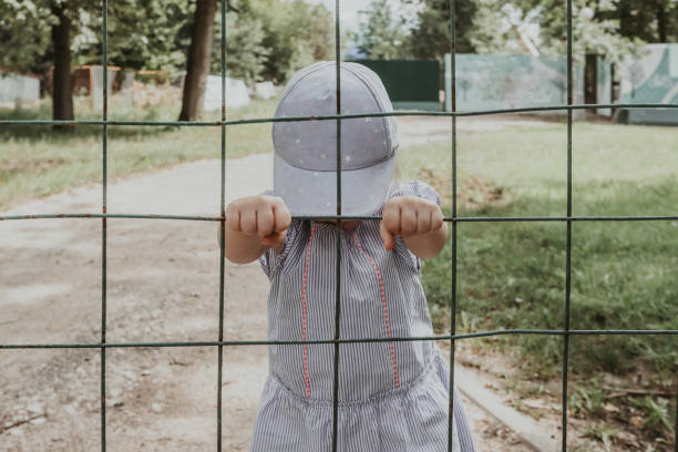 Little baby girl behind bars. The child holds onto the bars of the lattice. Little baby girl behind bars. The child stands with his head tilted and holds his hands on the bars of the lattice. The girl is dressed in a striped dress and a cap. Emotional stress and fear. Summer. girls in very short dresses stock pictures, royalty-free photos & images