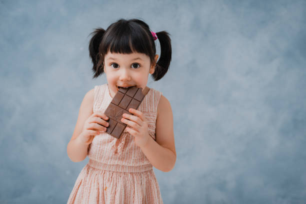 little baby girl eats chocolate and licks her finger on a grey blue Studio background. free space. stock photo