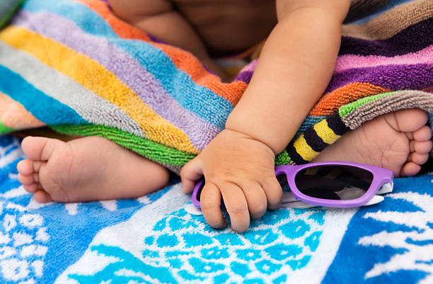 Little baby with barefeet sittting on a beach towel in the backyard and grabbing sunglasses stock photo