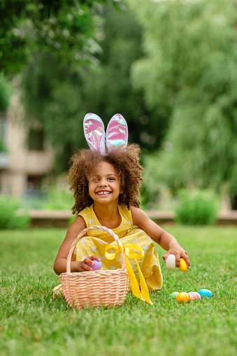Little Black girl with pink bunny ears gathering Easter colorful eggs during Easter egg hunt in garden