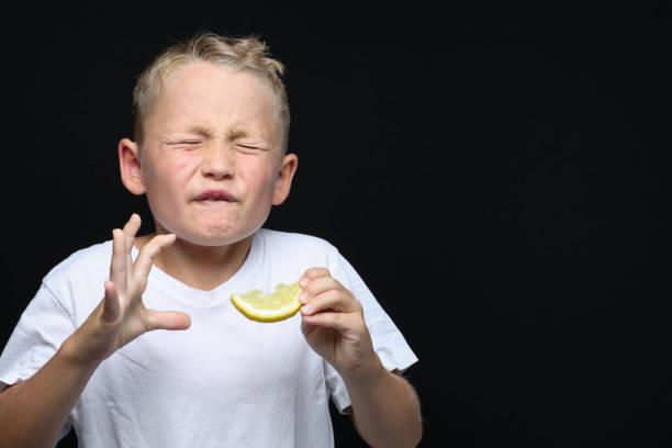 Little, blond boy is eating a piece of a lemon Little, blond boy is eating a piece of a lemon in front of black background and making a facial expression. sour taste stock pictures, royalty-free photos & images