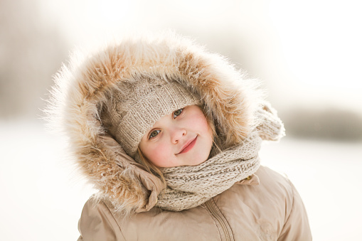 Little blond girl with rosy cheeks on a sunny frosty day. Beautiful winter portrait of a cute girl wearing beige fur hood, knit cap and scarf. Selective focus.