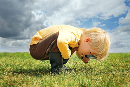 A little boy is exploring nature outside by looking at grass through a magnifying glass.