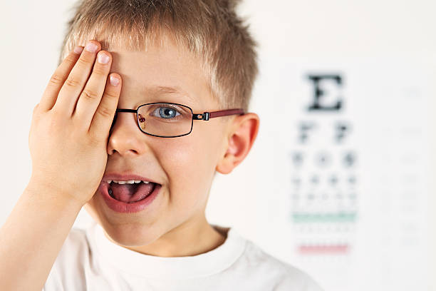 Little boy having eye exam. Eye examination of a smiling little boy wearing glasses. The boy is looking at the eye test chart and is saying what he sees. The boy is covering one eye. boys glasses stock pictures, royalty-free photos & images