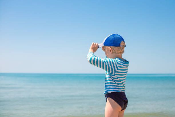 A little boy in a striped T-shirt and a blue cap stands on the seashore and looks into the distance. Happy summer days. copy space stock photo