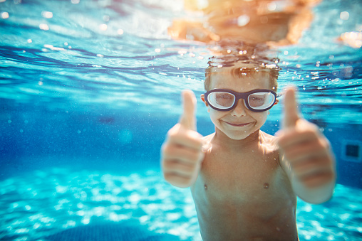 Little boy aged 6 swimming underwater. The boy is smiling at the camera showing thumbs up.\n