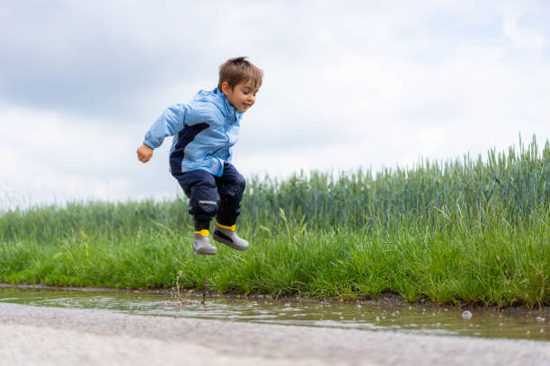 little boy jumping in water puddle four years old boy in rain clothes and wellies jumping in puddle on country road on bad weathers day boy jumping stock pictures, royalty-free photos & images