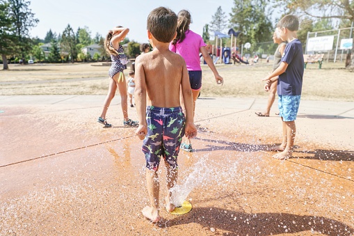Rear view of an mixed race elementary age boy playing in a fountain at the water park/ The child is cooling off and having fun playing outdoors with his older siblings and his cousins. A playground and homes are visible in the background.