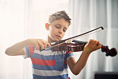 Closeup portrait of a focused little boy playing violin at home. The boy is aged 7 and is playing in a sunny room.
