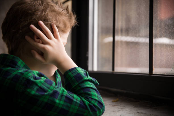 Little boy standing behind the window in sad mood. Sad Teenager looking in the Window and closing his ears with hands. Unhappy child in a plaid shirt. Alone at home. Upset. Little boy standing behind the window in sad mood. Sad Teenager looking in the Window and closing his ears with hands. Unhappy child in a plaid shirt. Alone at home. Upset. autism stock pictures, royalty-free photos & images