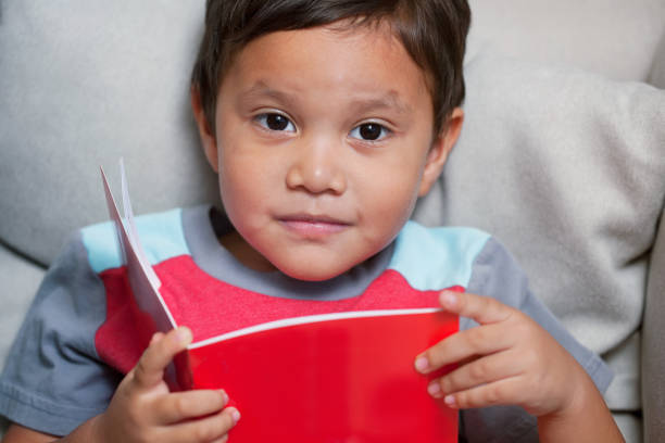 A little boy that is learning to read is holding a first reader type of book and has a thoughtful facial expression. stock photo