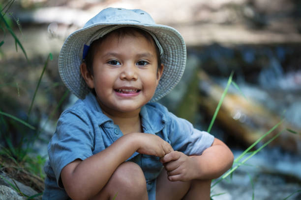 A little boy wearing his fishing hat, sitting next to the the rivers edge during summer. stock photo