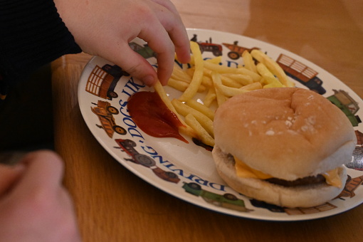 A little boys hand dipping oven cooked chips into tomato sauce, ketchup, on a child’s tractor plate with a hamburger and cheese in a bread roll