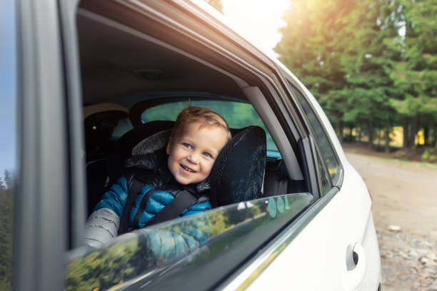 Little cute adorable happy caucasian toddler boy sitting in child safety seat car open window enjoy having fun making road trip nature countryside forest landscape. Family car travel by car concept stock photo