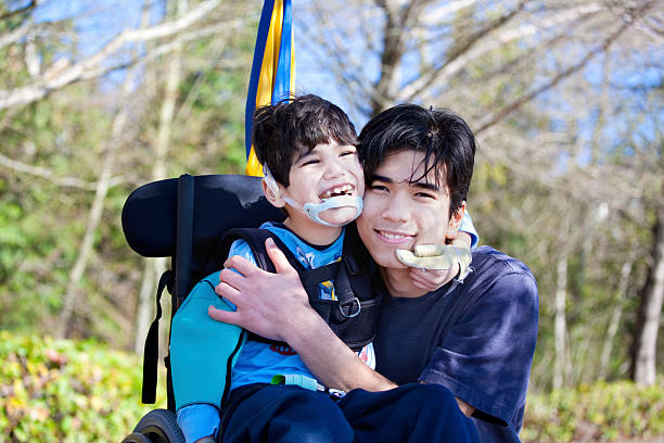 Little disabled boy in wheelchair hugging older brother outdoors stock photo