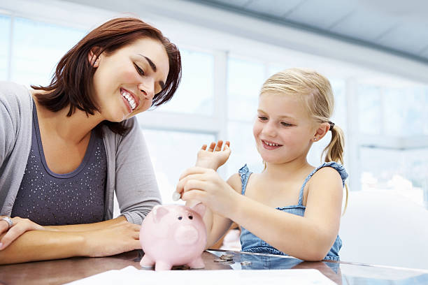 Little girl adding money to piggy bank Happy mother looking a her cute little daughter putting money in piggy bank allowance stock pictures, royalty-free photos & images