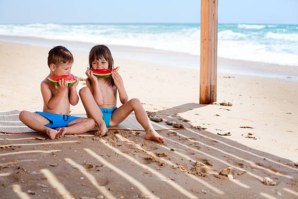 Little girl and boy enjoy eating watermelon on the beach Little girl and boy enjoy eating watermelon on the beach in the shade of big wooden parasol. Wonderful sand beach with the sendstones everywhere. little girls in bathing suits stock pictures, royalty-free photos & images