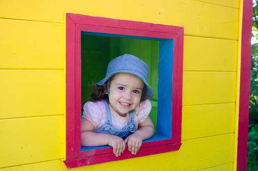 Little girl looking out of window from inside of colorful wooden toy house during summer day