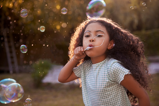 African-american cute little girl with curly hair blowing soap bubbles with heart-shaped hand-made wand in the garden during summer