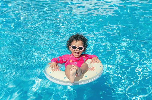 Little girl with pink glasses, curly hair and fuchsia swimsuit, enjoys the blue water pool with her round float on a sunny day
