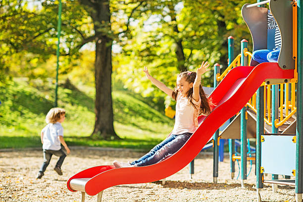 Little girl having fun while sliding. Children at the playground. Focus is on little girl sliding with her arms raised.   recess stock pictures, royalty-free photos & images